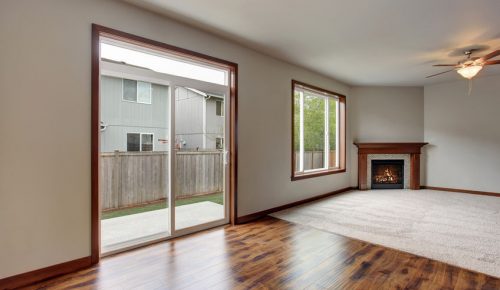 Large empty living room interior with carpet floor, fireplace and glass sliding doors leading to back yard.